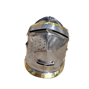 May include: A polished steel knight's helmet with brass detailing. The helmet has a visor with a horizontal slit and small ventilation holes. Brass bands adorn the brow and base, creating a historical aesthetic. The helmet is a classic piece of armour.