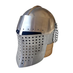Puede incluir: Un casco medieval plateado con visera y cadena adjunta. El casco tiene numerosos agujeros circulares pequeños en la visera y una base dorada. El casco está diseñado para proteger la cabeza y la cara.