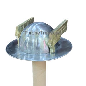 Könnte beinhalten: Ein silberfarbener Metallhelm mit zwei goldfarbenen Seitenflügeln. Der Helm hat einen breiten Rand und ist auf einem zylindrischen Ständer montiert. Der Text "Throne Treasure" ist auf dem Helm aufgedruckt.