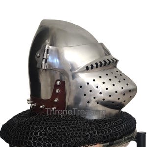 Puede incluir: Un yelmo de caballero plateado pulido con visera con pequeños orificios de ventilación. El yelmo tiene una visera con bisagras y está unido a una protección para el cuello de cota de malla negra. El texto "Throne Treasure" es visible.
