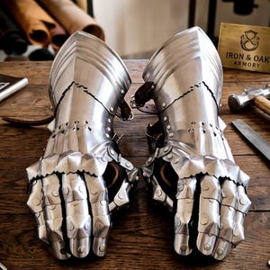 May include: A pair of polished steel gauntlets featuring articulated finger plates, riveted construction, and leather straps. A brass plaque nearby is engraved with the text IRON & OAK ARMORY. The metal armor pieces are designed to protect the hands and wrists.