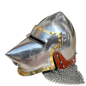 May include: A polished steel helmet with a pointed visor and brass detailing. The helmet has a riveted leather neck guard and a chainmail aventail. This medieval-style helmet is suitable for historical reenactment or display.