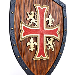 May include: A wooden shield featuring a dark brown textured surface with a black border accented by metal studs. A large red cross with gold trim is centered, flanked by two gold lion figures and two gold fleur-de-lis symbols on the wood panel.