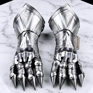Peut inclure: Paire de gantelets argentés ornés, avec des doigts articulés et des griffes pointues. Les gants métalliques présentent des gravures détaillées et sont exposés sur une surface blanche. Le texte "Est. 2024" est visible.