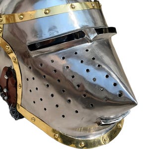 May include: A polished steel helmet with brass detailing. The helmet has a pointed visor with ventilation holes and a curved lower edge. Brass bands with rivets adorn the base and top. This helmet is likely for historical reenactment or display, reflecting medieval armour.