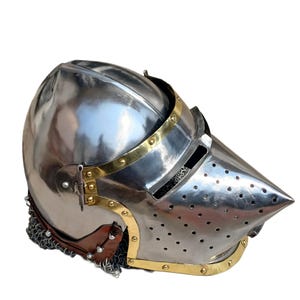 May include: A polished steel helmet with a pointed visor, brass detailing, and a chainmail neck guard. The helmet features a rounded crown and ventilation holes. The design is reminiscent of medieval armour.