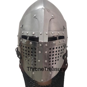 Pu&ograve; includere: Un elmo medievale in metallo color argento con una piastra frontale a forma di croce, fessure orizzontali per gli occhi e una sezione inferiore perforata per la ventilazione. L'elmo presenta rivetti visibili e una maglia di ferro nera.