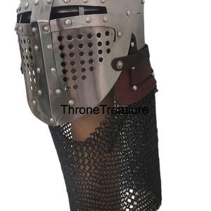 Pu&ograve; includere: Un elmo in stile medievale in metallo color argento con una piastra frontale rivettata dotata di una fessura orizzontale per gli occhi e una visiera perforata. L'elmo include un cinturino in pelle marrone scuro e un camaglio in maglia di ferro nero. Il testo ThroneTreasure &egrave; visibile.