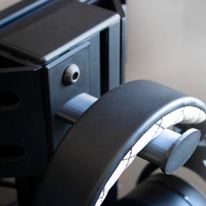 May include: Close-up of a black headphone stand with a curved arm and a textured pad. The stand is mounted on a black frame with a silver screw visible. The headphones have a white and black stitched design.