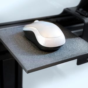 Op de afbeelding: Een zilveren en zwarte computermuis ligt op een getextureerde grijze plank die aan een zwart metalen frame is bevestigd. De muis heeft een glad, afgerond ontwerp en de plank lijkt gemaakt van een composietmateriaal. De opstelling suggereert een moderne werkplek.