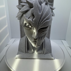 May include: A gray sculpture of a character's head with detailed facial features and a mask-like design on one side. The sculpture is on a white circular base, with a rectangular support behind the head.