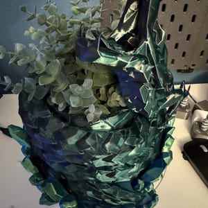 May include: A 3D-printed, faceted planter in shades of teal and blue, resembling a dragon's head. The planter is filled with faux eucalyptus greenery. The design features sharp angles and a textured surface, creating a unique decorative piece.