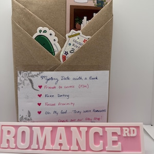 May include: A brown paper package with stickers and a bookmark. The bookmark has a bookshelf design. A pink sign that reads "ROMANCE RD" is in the foreground. A handwritten note lists romance novel tropes.