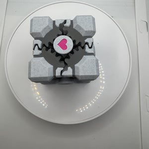 K&ouml;nnte beinhalten: Ein grau-wei&szlig;er W&uuml;rfel mit einem rosa Herzen in der Mitte. Der W&uuml;rfel hat ein strukturiertes Aussehen und ein zahnrad&auml;hnliches Design um das Herz. Der W&uuml;rfel befindet sich auf einer wei&szlig;en, kreisf&ouml;rmigen Plattform.