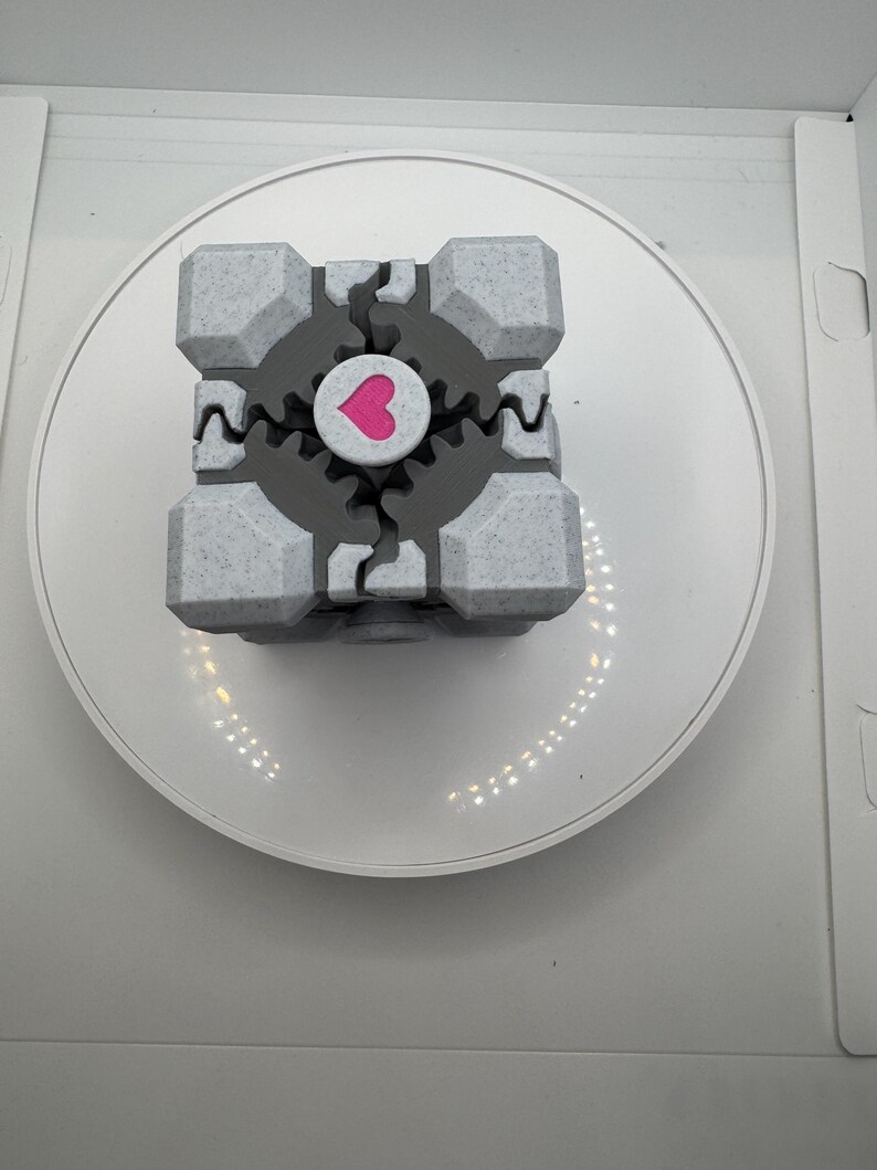 K&ouml;nnte beinhalten: Ein grau-wei&szlig;er W&uuml;rfel mit einem rosa Herzen in der Mitte. Der W&uuml;rfel hat ein strukturiertes Aussehen und ein zahnrad&auml;hnliches Design um das Herz. Der W&uuml;rfel befindet sich auf einer wei&szlig;en, kreisf&ouml;rmigen Plattform.