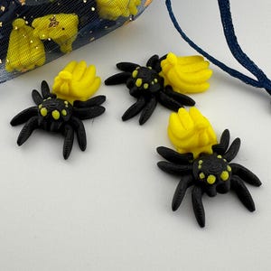 May include: Black spider-shaped embellishments with yellow accents and a yellow flower-like shape. A blue mesh bag with gold stars and yellow shapes is in the background. The spiders have eight legs and are on a white surface.