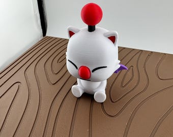 May include: A white 3D printed figurine of a cat-like creature with a red nose, red pom-pom antenna, and small purple wings. The figure has closed eyes, pointed ears, and is in a seated position with its legs extended forward on a brown surface.