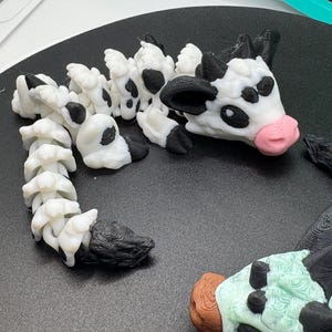May include: A 3D-printed cow figurine with a white body, black spots, and a pink nose. The cow's head and body are segmented, and the figurine is lying on a black surface. The cow has black horns and ears.