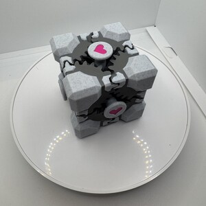 K&ouml;nnte beinhalten: Ein grau-wei&szlig;er W&uuml;rfel mit Zahnradmuster und rosa Herzakzenten. Der W&uuml;rfel steht auf einer wei&szlig;en Oberfl&auml;che. Der W&uuml;rfel hat ein geometrisches Design und ist wahrscheinlich ein Dekorationsartikel oder ein Spielzeug.