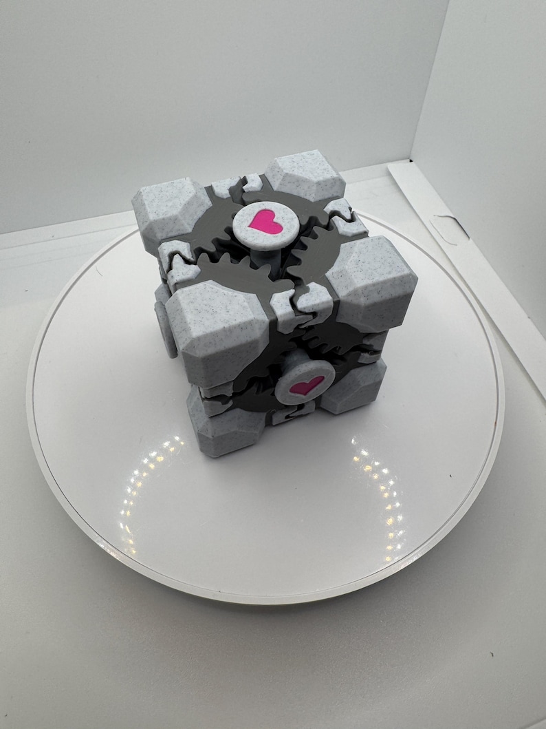 K&ouml;nnte beinhalten: Ein grau-wei&szlig;er W&uuml;rfel mit Zahnradmuster und rosa Herzakzenten. Der W&uuml;rfel steht auf einer wei&szlig;en Oberfl&auml;che. Der W&uuml;rfel hat ein geometrisches Design und ist wahrscheinlich ein Dekorationsartikel oder ein Spielzeug.