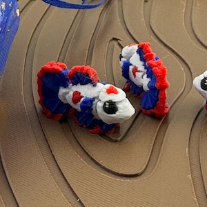 May include: A pair of small, decorative fish figurines. The fish are white with red and blue accents, and have detailed fins. They are placed on a brown, textured surface, with a blue ribbon visible in the background.