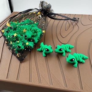 May include: A collection of small, green lizard figurines with black eyes. Some are in a black mesh bag decorated with gold stars and moons. The figurines are displayed on a brown, textured surface.