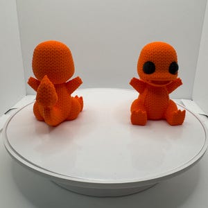 May include: Two orange crochet figures of a Charmander, a Pokémon character, are displayed. The figures have black eyes and are posed with their arms outstretched. The figures are sitting on a white surface.