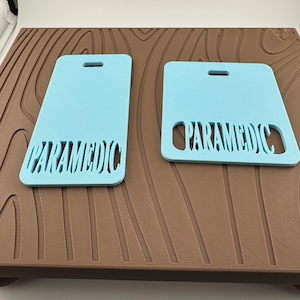 May include: Two light blue rectangular tags with rounded corners, each featuring a horizontal slot at the top and the word PARAMEDIC cut out in a bold, stylized font at the bottom. One tag is taller and rectangular, while the other is square.