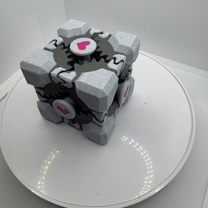 K&ouml;nnte beinhalten: Ein grau-wei&szlig;er W&uuml;rfel mit strukturierter Oberfl&auml;che, schwarzen zahnradartigen Details und rosa Herzakzenten. Der W&uuml;rfel ist ein geometrisches Design, m&ouml;glicherweise ein Spielzeug oder Dekorationsartikel, vor wei&szlig;em Hintergrund.