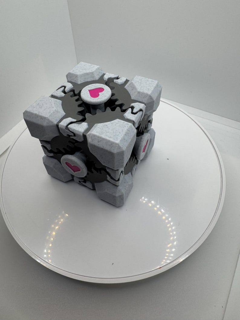 K&ouml;nnte beinhalten: Ein grau-wei&szlig;er W&uuml;rfel mit strukturierter Oberfl&auml;che, schwarzen zahnradartigen Details und rosa Herzakzenten. Der W&uuml;rfel ist ein geometrisches Design, m&ouml;glicherweise ein Spielzeug oder Dekorationsartikel, vor wei&szlig;em Hintergrund.