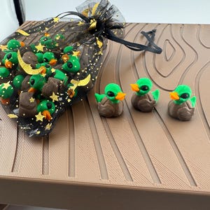May include: A black mesh bag with gold stars and a drawstring, filled with small, brown and green duck figurines. Three duck figurines are arranged on a brown surface. The ducks have green heads, orange beaks, and brown bodies.