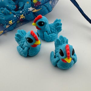 May include: Three blue chicken figurines with red combs and yellow beaks are displayed. The chickens are in a playful pose, with their wings slightly spread. A blue mesh bag with gold stars is in the background.