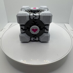 K&ouml;nnte beinhalten: Ein grau-schwarzer W&uuml;rfel mit einem Herzdesign. Der W&uuml;rfel hat eine strukturierte Oberfl&auml;che und eine schwarze Mitte mit einem rosa Herzen. Die Ecken des W&uuml;rfels sind grau und er steht auf einer wei&szlig;en Oberfl&auml;che.