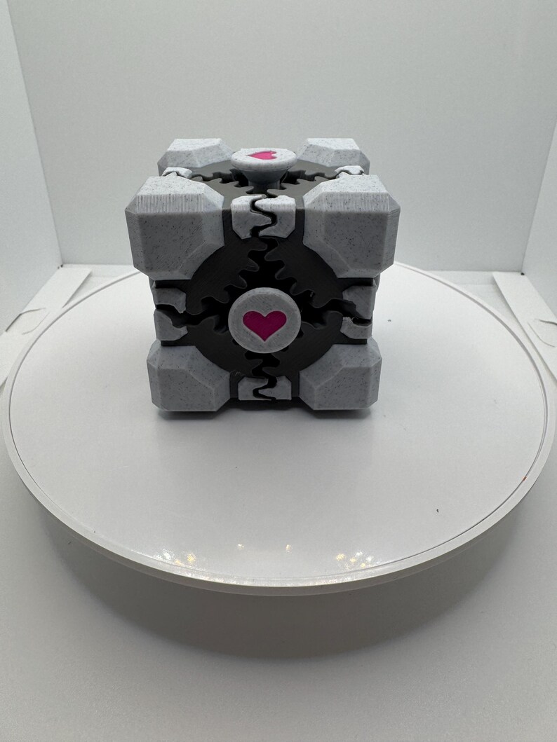 K&ouml;nnte beinhalten: Ein grau-schwarzer W&uuml;rfel mit einem Herzdesign. Der W&uuml;rfel hat eine strukturierte Oberfl&auml;che und eine schwarze Mitte mit einem rosa Herzen. Die Ecken des W&uuml;rfels sind grau und er steht auf einer wei&szlig;en Oberfl&auml;che.