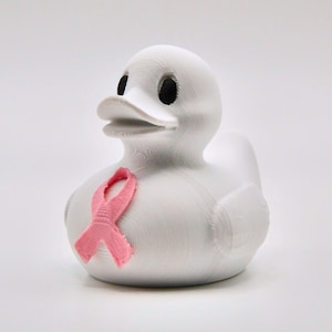 May include: A white rubber duck with black eyes and a pink ribbon, symbolizing breast cancer awareness. The duck is a classic toy, often used in bathtubs or as a decorative item. The pink ribbon is a symbol of support.