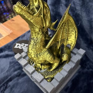 May include: A golden dragon statue with its mouth open, wings spread, and perched on a gray castle-like base. The dragon is detailed with textured scales. The base has a D&D logo.