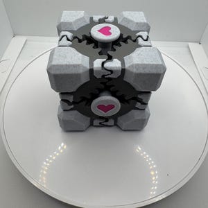 K&ouml;nnte beinhalten: Grau-schwarzer W&uuml;rfel mit einem rosa Herz auf jeder Seite. Der W&uuml;rfel hat ein zahnrad&auml;hnliches Design und steht auf einer wei&szlig;en Oberfl&auml;che. Dieser 3D-gedruckte Artikel ist ein einzigartiges Dekorationsobjekt.