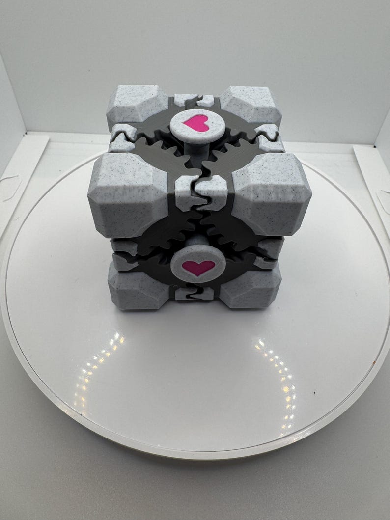 K&ouml;nnte beinhalten: Grau-schwarzer W&uuml;rfel mit einem rosa Herz auf jeder Seite. Der W&uuml;rfel hat ein zahnrad&auml;hnliches Design und steht auf einer wei&szlig;en Oberfl&auml;che. Dieser 3D-gedruckte Artikel ist ein einzigartiges Dekorationsobjekt.