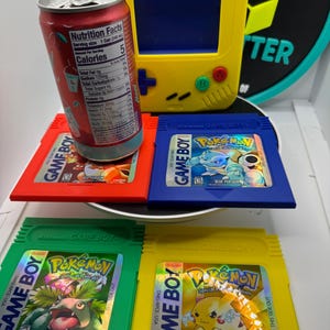 May include: A yellow Game Boy handheld console with a blue screen, red, green, and blue Pokemon Game Boy cartridges, and a red can of a beverage. The cartridges are labeled "Pokemon" and "Game Boy".