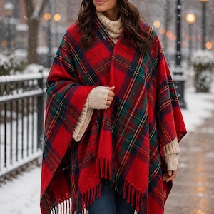 May include: A red plaid poncho with green and navy accents and fringe trim. The poncho is worn with a cream-colored knit hat and sweater. The person is wearing blue jeans and brown boots. The background is a snowy outdoor scene.