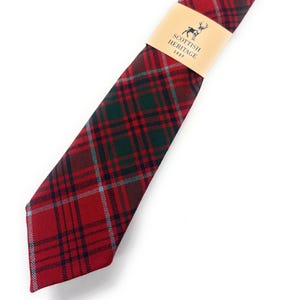 May include: A red, green, and navy tartan tie with a light blue accent. The tie is wrapped with a tan paper band that reads "Scottish Heritage Shop" with a deer logo. The tie is angled diagonally.