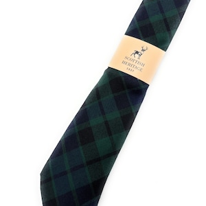 Puede incluir: Una corbata a cuadros verdes y azul marino, con una banda de papel que dice "Scottish Heritage Shop" y un logotipo de ciervo. La corbata está colocada en diagonal, mostrando el diseño de tartán clásico.