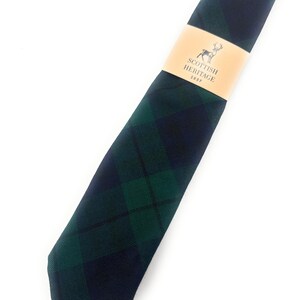 Puede incluir: Una corbata a cuadros verdes y azul marino con una banda de papel color canela que dice "Scottish Heritage Shop" con un logotipo de ciervo. La corbata está en ángulo diagonalmente sobre un fondo blanco.
