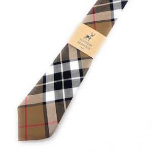 May include: A tartan necktie in shades of tan, black, white, and red. The tie features a brown paper band with the text "Scottish Heritage Shop" and a deer logo. The tie is displayed on a white background.