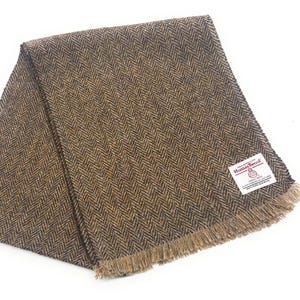May include: A brown and black herringbone tweed scarf with a fringed edge. The scarf is folded and has a label that reads "Harris Tweed". The scarf is made of wool.