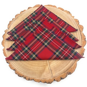 May include: Four red tartan napkins with green, yellow, and white detailing are displayed on a wooden slice. The napkins are folded into triangles, highlighting the traditional plaid design. Perfect for a festive dinner setting.