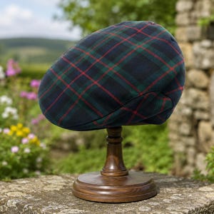 May include: A flat cap featuring a dark blue, green, and red plaid tartan pattern. The cap is displayed on a dark brown wooden stand. The fabric has a smooth texture and a rounded, structured shape that tapers toward the front brim.