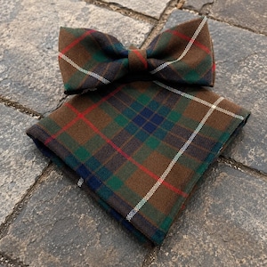 May include: A bow tie and a matching pocket square featuring a brown, green, blue, and red plaid tartan pattern with thin white diagonal lines. The set is composed of a pre-tied bow tie and a folded square fabric piece with a textured appearance.