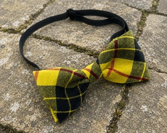 May include: A pre-tied bow tie featuring a vibrant yellow, black, and thin red plaid pattern. The bow tie is constructed from a textured fabric and includes an adjustable black elastic neck strap with a plastic slider for a secure fit.