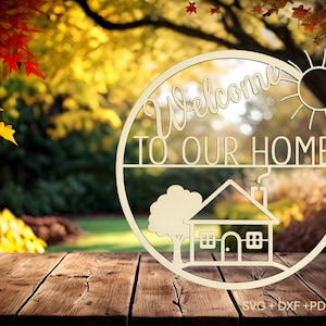 May include: A wooden sign with the words "Welcome TO OUR HOME" cut out in a circular design. The sign features a house, tree, and sun illustration. The background is a blurred autumn scene.